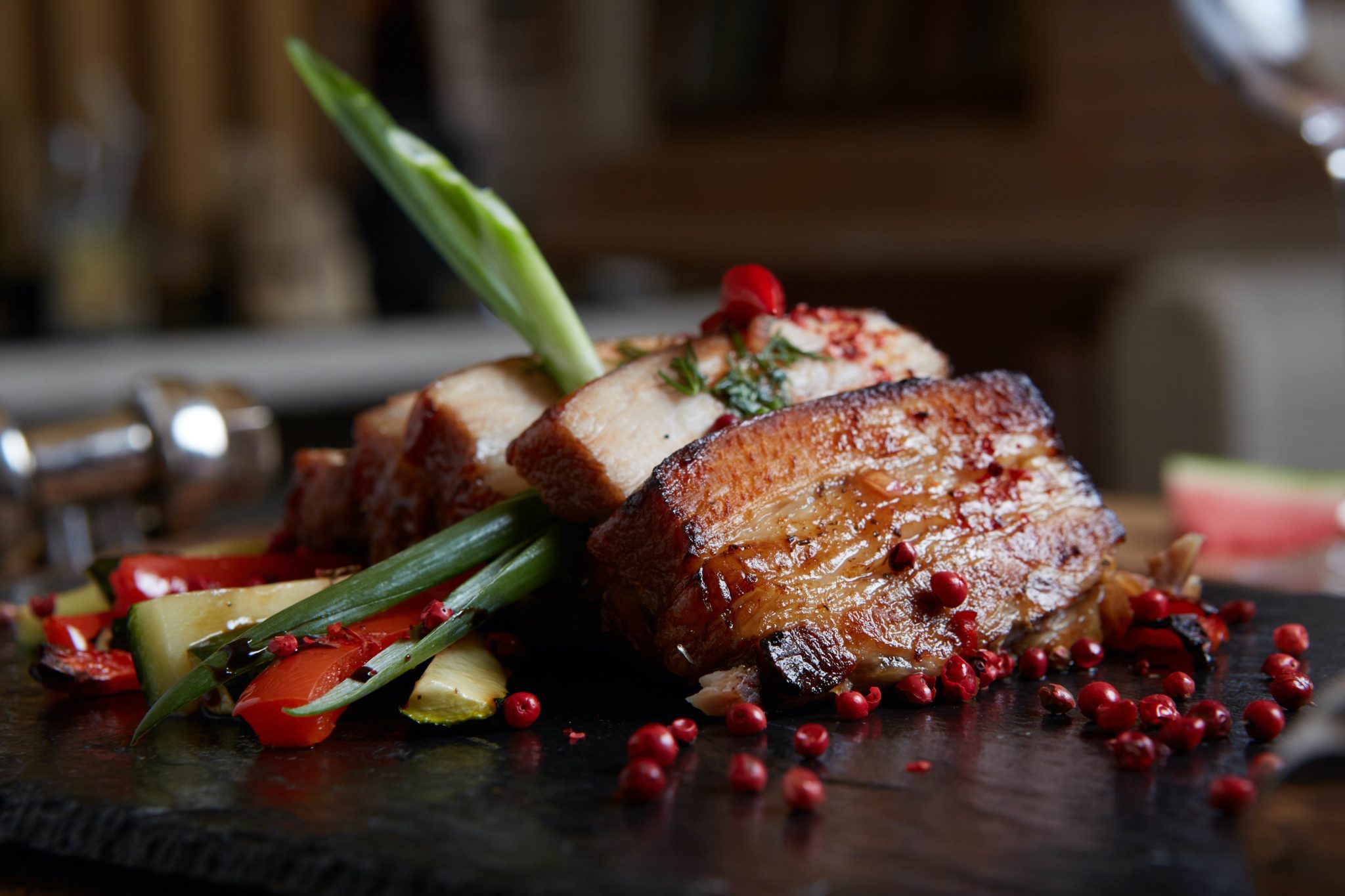 AdapTable Meals Close-up of glazed pork belly, garnished with green onions and peppercorns, with sliced red bell peppers and cucumbers.